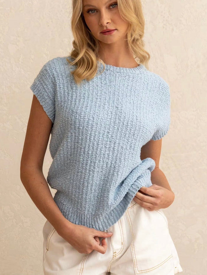 Paper Heart Top. A woman wearing a light blue short-sleeve knit top with a textured waffle pattern, paired with cream shorts, set against a neutral beige textured wall.