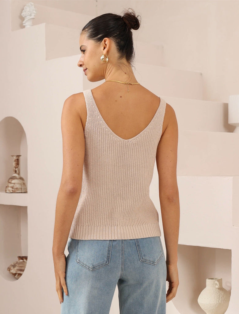 Woman wearing a beige knitted tank top with scooped neckline and blue jeans in a minimalistic room.